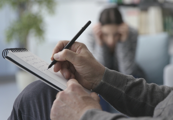 A psychologist takes the anamnesis of a patient preparing the treatment for depression in Dubai. The picture is showing a person sitting in the chair taking notes in a notebook. There is a blurred silhouette of a patient in the background.