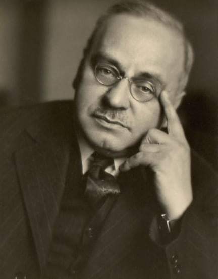 An old fashioned black on white picture of Dr. Alfred Adler. In the picture he is wearing round metal glasses and is sporting a mustache. His head is tilted to the side and he is pointing his index finger to his temple. He is wearing a dark suit, vest, and tie over a white shirt. This image reflects Adler's biography.
