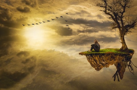 feeling depressed in Dubai. A cloudy sky with birds flying in formation on the top left, and the sun shining through below the birds. On the right side of the picture a small piece of land with a naked tree, grass, and visible tree roots. A person sitting alone in front of the tree. Feeling depressed can make people feel lonely and alone in the world.